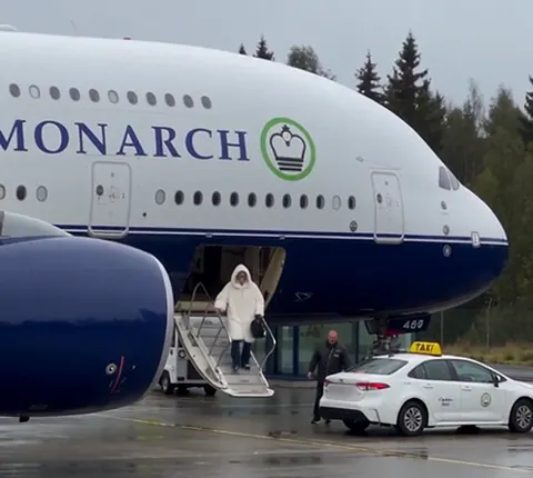 Sinivalkoinen jättimäinen matkustajakone, jonka kyljessä lukee MONARCH, seisoo lentoasemalla, ja portaita laskeutuu valkoiseen turkkiin pukeutunut mies kohti taksia.