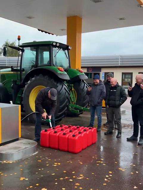 Traktori tankkaa ABC-huoltoasemalla, mies täyttää punaisia kanistereita ja ihmiset kuvaavat puhelimillaan.