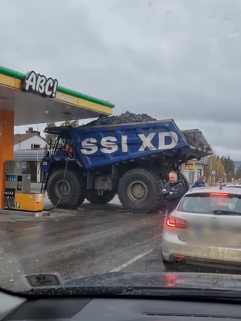 Jättimäinen sininen kaivosauto ABC-aseman katoksen vieressä, ja tavallisia autoja jonottaa sen edessä.