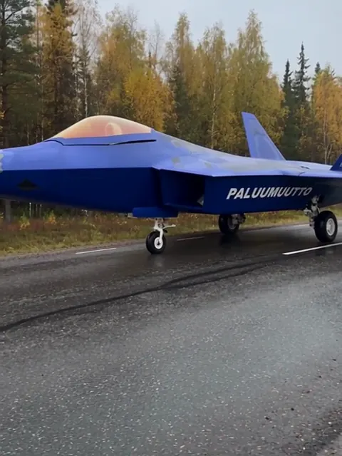 Sininen hävittäjä seisoo keskellä Sastamalan valtatietä, kyljessä teksti PALUUMUUTTO ja taustalla syksyinen metsä.