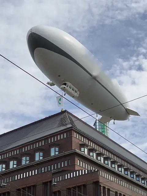 Valkoinen ilmalaiva leijuu punatiilisen tavaratalon yllä Helsingin keskustassa.