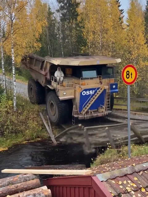 Valtava kaivosauto on kaatunut keula edellä puroon murtuneen puusillan kohdalla, ja vieressä on kahdeksan tonnin painorajoituskyltti.