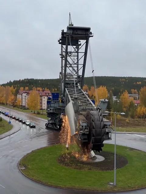 Jättimäinen kauhapyöräkaivuri jauhaa liikenneympyrän veistosta kipinöiden lentäessä.