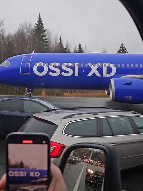Sininen matkustajakone, jonka kyljessä lukee OSSI XD, ohittaa autojonon tien pientareella. Kuva otettu auton ikkunasta.