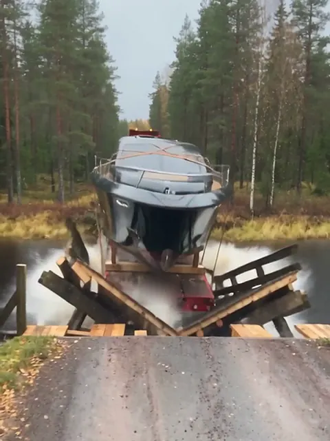 Superjahti lavetin kyydissä keskellä katkennutta puusiltaa, ja perävaunu on painunut metsäpuroon.
