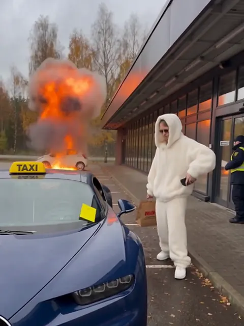 Sininen Bugatti rekisterikilvellä DUE-1 pysäköintialueella, taustalla liekehtivä auto ja valkoiseen turkkiin pukeutunut mies.