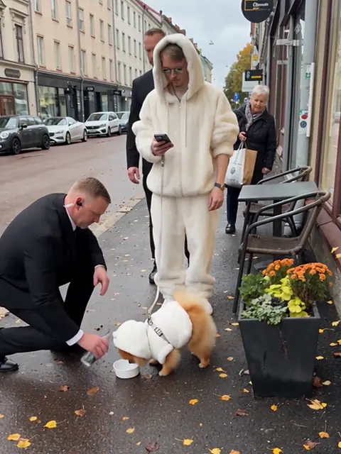 Valkoiseen turkkiin pukeutunut mies katsoo puhelintaan, kun pukumies antaa valkoiseen takkiin puetulle pomeranianille vettä kadulla.