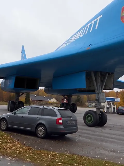 Sinisen pommikoneen siipi huoltoaseman pihalla aivan pysäköidyn harmaan farmariauton yläpuolella.