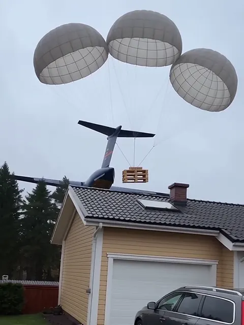 Kolme laskuvarjoa laskee lavaa omakotitalon pihalle, taustalla matalalla lentävä rahtikone.