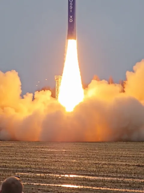 Sininen raketti nousee liekkien ja savun saattelemana sastamalalaiselta pellolta, etualalla kuvaaja aidan takana.