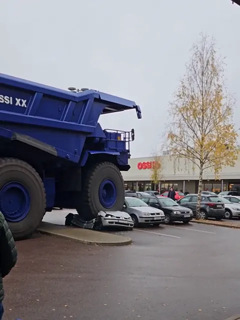 Sininen kaivosauto seisoo litistyneen henkilöauton päällä kauppakeskuksen parkkipaikalla.