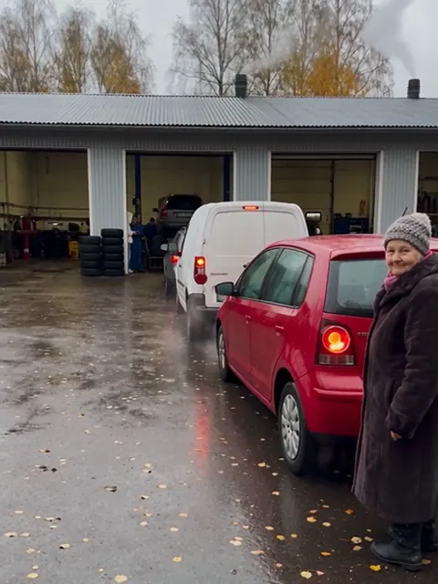 Eläkeläinen seisoo rengasliikkeen pihalla autojonon perässä, taustalla huoltohalli ja rengaspinoja.