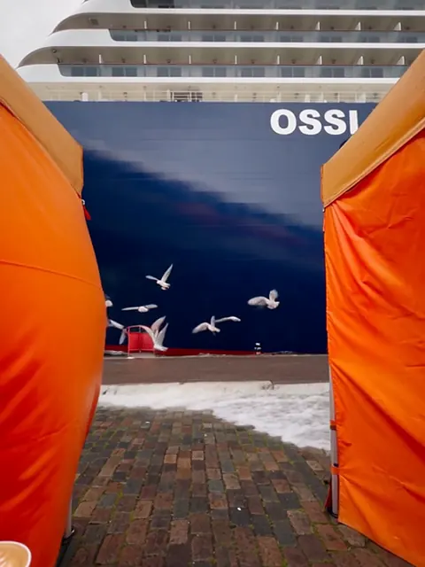 Valtava risteilyalus on kiinnittynyt Kauppatorin laituriin oranssien torikojujen taakse, ja etualalla on munkkeja.