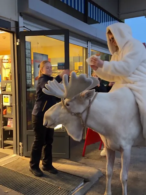 Valkoiseen turkkiin pukeutunut mies istuu valkoisen hirven selässä R-kioskin ovella, myyjä seisoo vieressä.