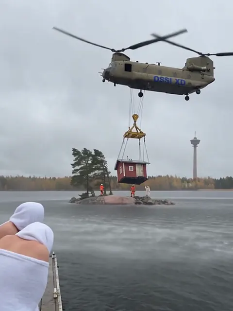 Raskas helikopteri kantaa punaista saunamökkiä ketjuissa Näsijärven yllä, ja laiturilla seisoo pyyhkeeseen kääriytynyt saunoja.